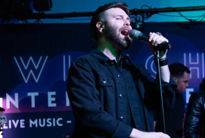 Bearded male singer in a black shirt performing on stage under blue lighting.