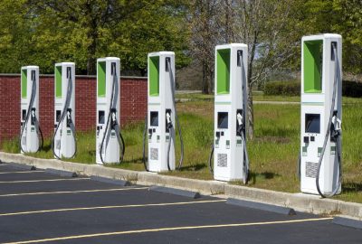 *   Image: Six white electric vehicle (EV) charging stations lined up in a parking lot.
    *   Details: White stations with