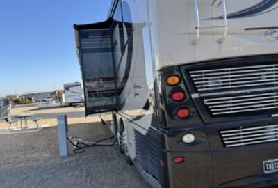 Luxury RV with an extended slide-out, parked on gravel and connected to a power pedestal.