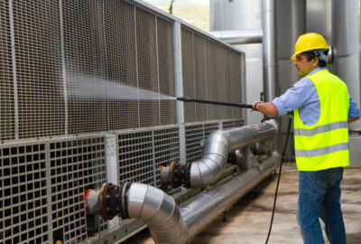 Worker in a yellow hard hat and safety vest using a pressure washer to clean industrial cooling equipment.
