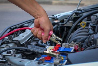 *   Image content: A person's hand is holding a red jumper cable clamp, attempting to attach it to a car battery under the ho