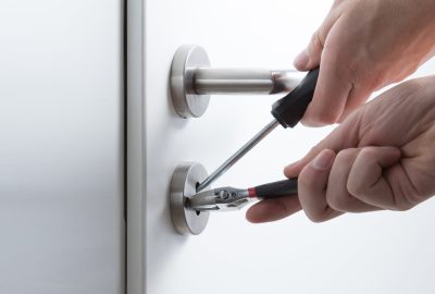 Hands using a screwdriver and pliers to force open a door lock.
