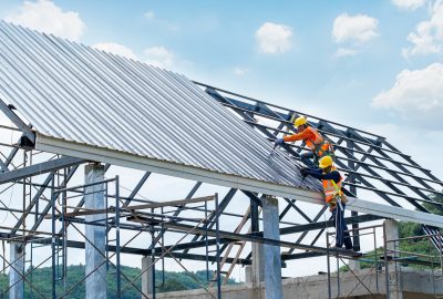Construction workers in safety gear installing corrugated metal roofing on a steel structure.