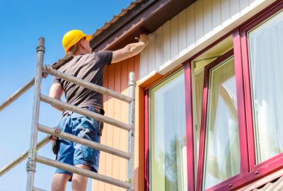 Worker in a yellow hard hat standing on scaffolding to paint the exterior trim of a house white.