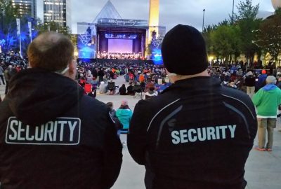 Two security guards stand with their backs to the camera, overseeing an outdoor orchestral concert in a city plaza.