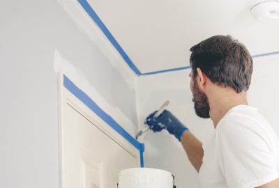 A man wearing a blue glove uses a paintbrush to paint a white wall with blue painter's tape along the edges.