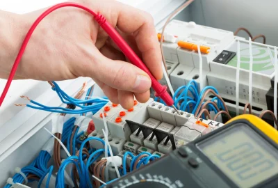 A hand using a multimeter probe to test the voltage on a terminal block in an electrical panel.