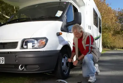 Man using a lug wrench to change the front tire of a white RV parked on a roadside.
