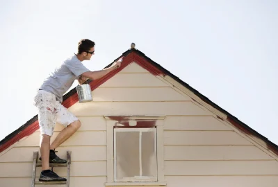 Man standing on a ladder painting the exterior roof trim of a house red.