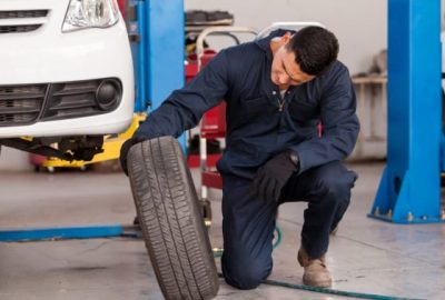 *   Image content: A mechanic in blue overalls and gloves is kneeling in a garage, inspecting a car tire. A white car is rais