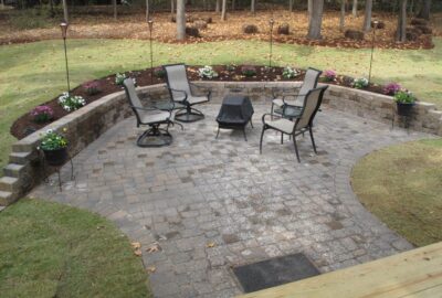 Stone patio with four outdoor chairs around a covered fire pit, enclosed by a curved stone wall and garden beds.