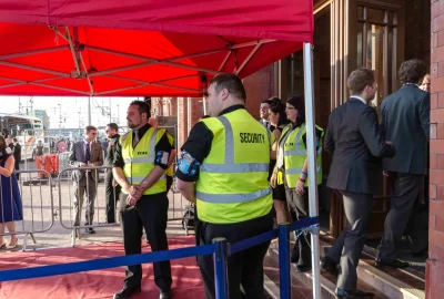 Security personnel in neon yellow vests monitor a red carpet event entrance under a red canopy.