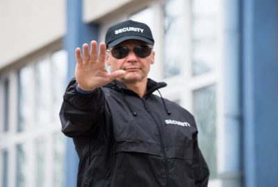Security guard in a black uniform and sunglasses making a stop gesture.
