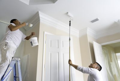 Two painters in white uniforms painting a ceiling and crown molding with a paintbrush on a ladder and a paint roller.