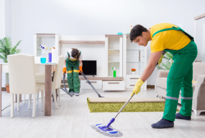 Two professional cleaners in green overalls mopping and vacuuming a modern living room.