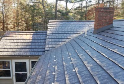 High-angle view of a house with a dark grey slate shingle roof, a red brick chimney, and trees in the background.
