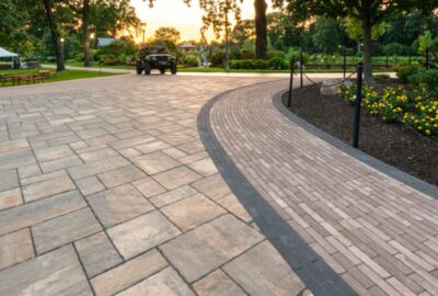 Landscaped park plaza with large stone pavers and a curved brick border, surrounded by trees and gardens at sunset.