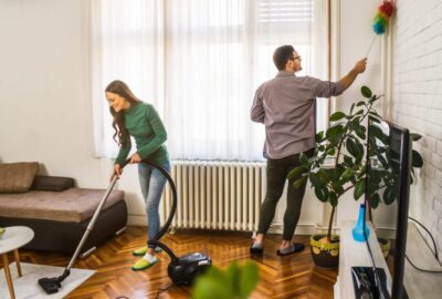 Couple cleaning a living room: a woman vacuums the floor while a man dusts the upper wall.