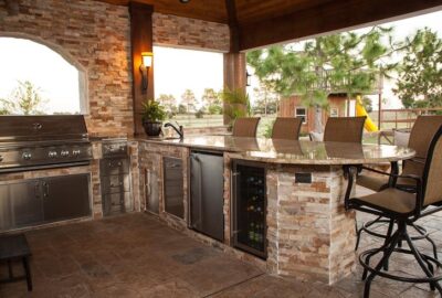 Outdoor kitchen with a built-in stainless steel grill, stone counters, bar seating, and a wine fridge under a covered patio.