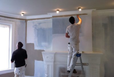 Two painters renovate a room, one on a stepladder painting near the ceiling above a covered fireplace.