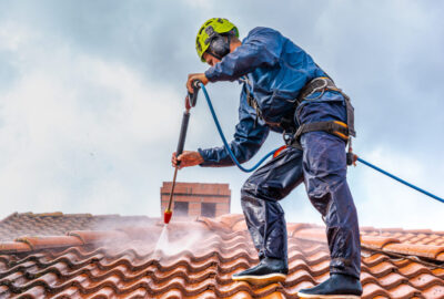 Worker in protective gear and a safety harness pressure washing a red tiled roof.