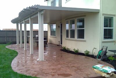 A large, curved stamped concrete patio with a white pergola structure attached to a two-story house.