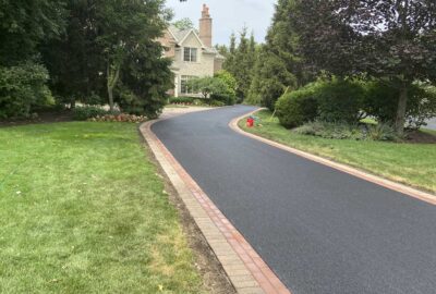 A newly paved black asphalt driveway with a brick border winding through a green lawn toward a large house.