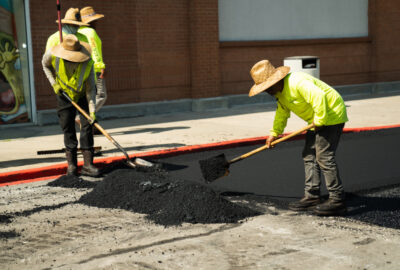 Workers in high-visibility yellow shirts and straw hats shoveling black asphalt on a road.