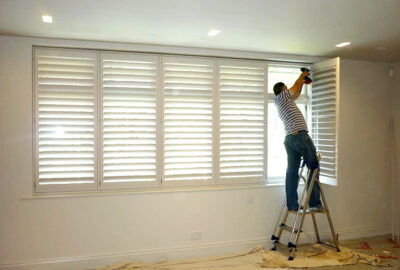 A man on a ladder uses a power drill to install white plantation shutters on a large window.