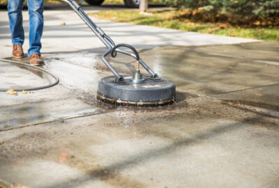 Person using a circular surface cleaner attachment to pressure wash a concrete driveway.