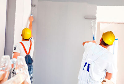 Two painters in yellow hard hats painting an interior wall light grey with rollers.