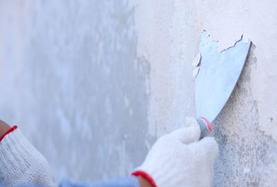 Person in work gloves using a putty knife to scrape old, peeling paint off a wall.