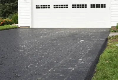 Freshly sealed black asphalt driveway leading to a white garage door, flanked by green grass and flowers.
