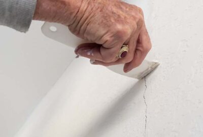 A person uses a white putty knife to apply filler to a crack in a white textured wall.