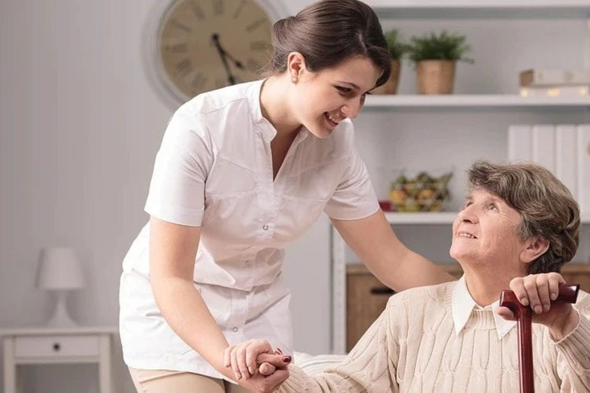 *   Image: A young woman in a white caregiver/nurse uniform is smiling and holding the hand of an elderly woman. The elderly