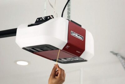 A hand pulls the emergency release cord on a white and red LiftMaster garage door opener mounted to a ceiling.