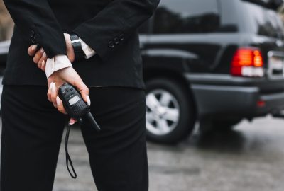 A person in a black business suit stands with their hands behind their back, holding a small two-way radio.
