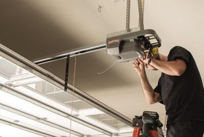 A man in a black t-shirt repairs or installs a garage door opener motor mounted to the ceiling.