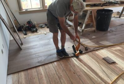 A man installs wide-plank hardwood flooring using a floor tapping block and mallet in a room under construction.