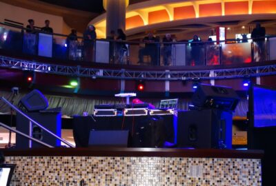 DJ equipment, speakers, and a laptop on a stage in a dimly lit nightclub with people watching from a balcony above.