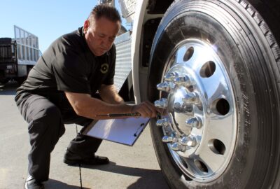 Man inspecting a semi-truck's large chrome wheel while writing on a clipboard.