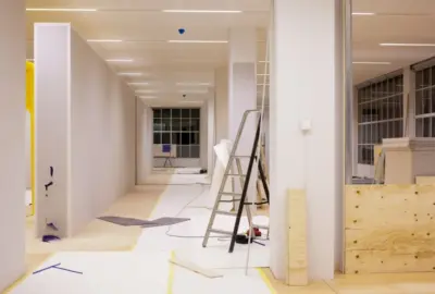 An empty, brightly lit office space under construction with drywall, a ladder, and various building materials.