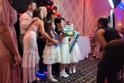 People and children in formal attire gathered near a red, black, and gold balloon arch at a celebration.
