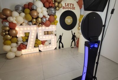 Disco-themed 75th birthday party setup with a balloon arch, light-up "75" numbers, and photo booth equipment.