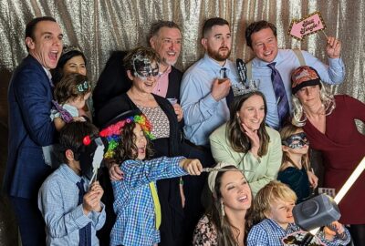 A joyful group of adults and children posing with photo booth props against a silver sequined backdrop.