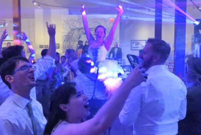 Guests cheer and dance under blue lights at a wedding celebration, with the bride lifted high in the air.
