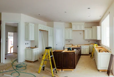 Kitchen under construction with white and dark wood cabinets, yellow step ladders, and unfinished floors.