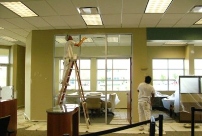 Two painters in white clothing painting an office interior with furniture protected by plastic sheets.
