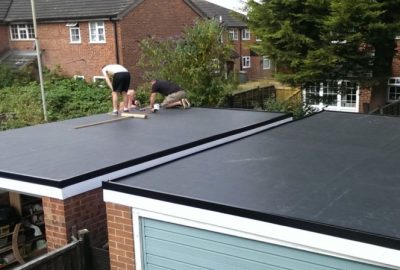 Two men installing a new black rubberized flat roof on a brick garage.