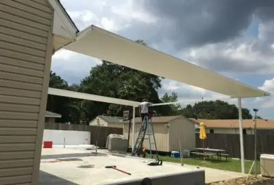 A man on a ladder installs a large white metal patio cover over a concrete pad in a backyard.